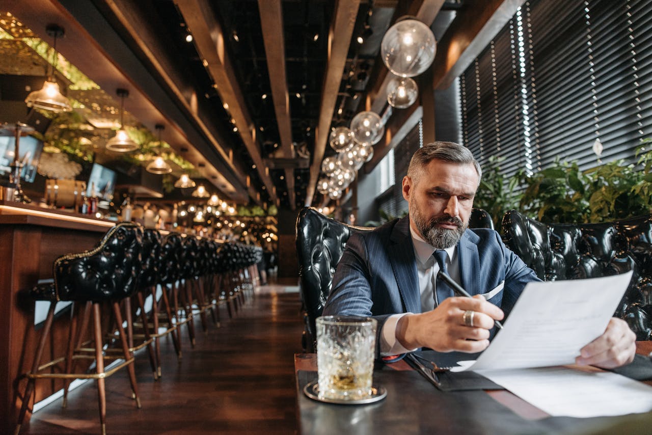 Stylish businessman in suit reviews documents at a modern bar with whiskey glass in view.