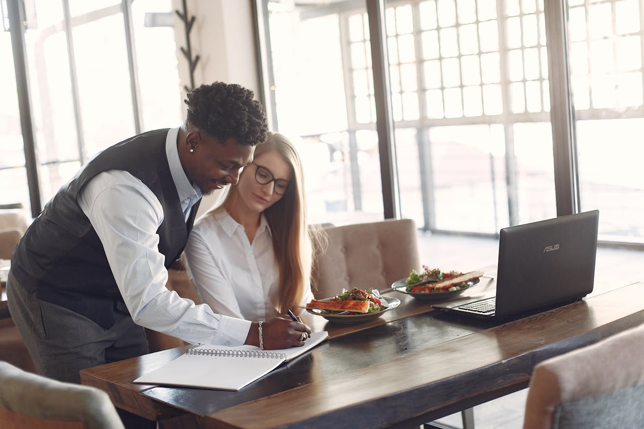 Professional colleagues discussing a project at a café over lunch, using a laptop.